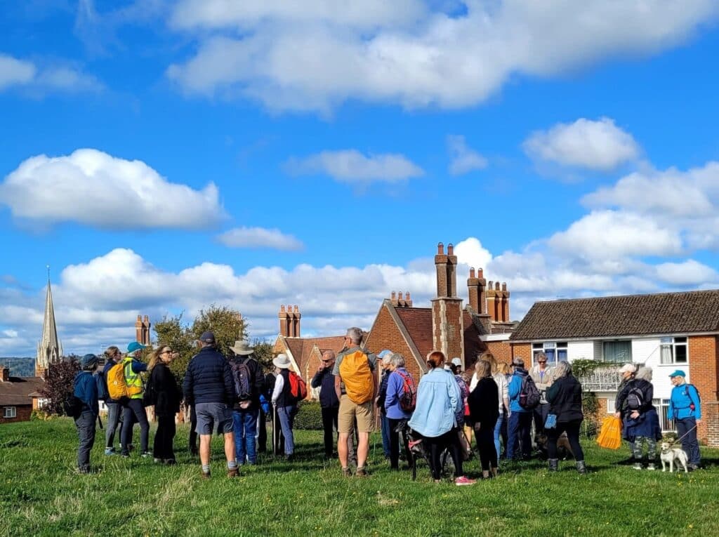 Group of walkers on a grassy hill with Dorking rooftops and a church spire under a bright blue sky.